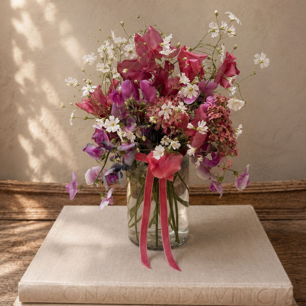A mixed wildflower bouquet in a clear glass jar vase, tied with a pink ribbon, displayed on a beige book atop a rustic wooden surface against a textured beige wall.